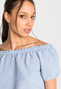 Blusa com ombros à mostra em azul claro e riscas brancas. Apresenta decote elástico e mangas curtas bufantes com textura enrugada.