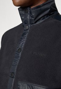 Mørkegrå jakke med en myk fleecekropp, skinnende nylon-detaljer, høy krage, trykknapper og diskret logo.