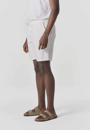 Person trägt weiße Shorts mit Kordelzug und beige Sandalen mit doppeltem Riemen, steht vor einem schlichten hellen Hintergrund.