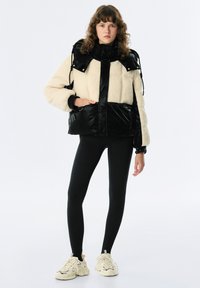 Czarna i kremowa kurtka z polarem, o błyszczącej powierzchni, w oversize'owym kroju, z wysokim kołnierzem i regulowanymi paskami. Połączona z czarnymi legginsami i butami.
