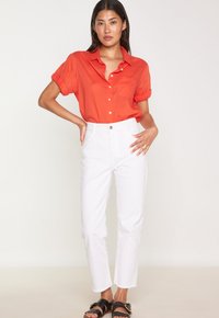 Camicia rossa a maniche corte con bottoni e taschino sul petto, abbinata a pantaloni bianchi a vita alta con gamba dritta e orli sfilacciati, e sandali neri.