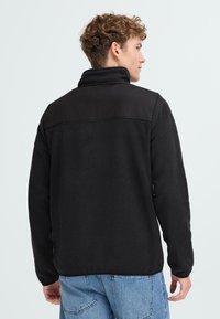 Chaqueta de forro polar negra con cuello alto, sección superior en contraste y mangas largas. Textura suave con un ajuste relajado.