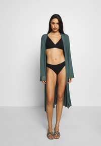 Dolga, zelena kimono obleka z čipkastimi detajli preko črnega bikinija. Model nosi natikače s potiskom leopardje kože. Gladka tekstura in sproščen kroj.