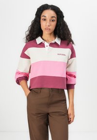Kvinne med langt krøllet hår iført en kort, stripete rugbytrøye med "RIP CURL"-logo og brune bukser, stående med den ene hånden i lommen.