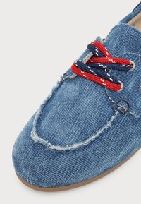 Chaussure décontractée en denim avec des bords effilochés et des lacets rouges et bleu marine passant par des œillets en métal sur un fond clair.