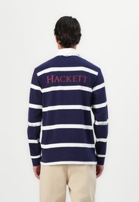 Polo à manches longues rayé bleu marine et blanc avec un col blanc, portant "HACKETT" imprimé en rouge au dos. Tissu en coton lisse.