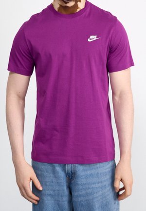 Mann trägt ein lila Nike T-Shirt und blaue Jeans, steht vor einem schlichten weißen Hintergrund mit entspannten Armen an seiner Seite.