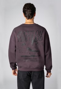 Dunkelvioletter Pullover mit Grafikdruck auf der Rückseite, der den Text „KONZENTRIERE DICH AUF DAS GUTE“ und schwarze Cartoonfiguren zeigt.