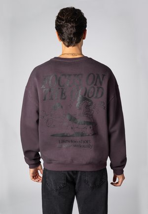 Dunkelvioletter Pullover mit Grafikdruck auf der Rückseite, der den Text „KONZENTRIERE DICH AUF DAS GUTE“ und schwarze Cartoonfiguren zeigt.