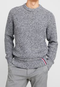 Grauer, strukturierten Strickpullover mit Rundhalsausschnitt, gerippten Bündchen und Saum. Besitzt einen kleinen roten, weißen und blauen Akzent am Saum.