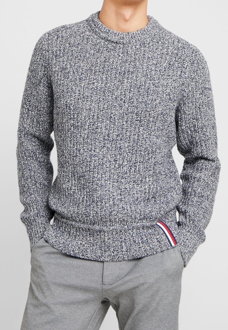 Grauer, strukturierten Strickpullover mit Rundhalsausschnitt, gerippten Bündchen und Saum. Besitzt einen kleinen roten, weißen und blauen Akzent am Saum.