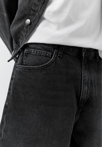 Nærbillede af sorte denimjeans med forlomme og hvid skjorte, der er stoppet ind, kombineret med sort denimjakke.