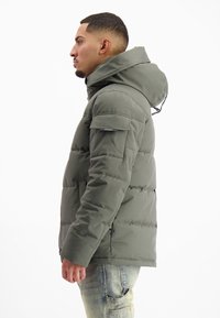 Groene gewatteerde jas met een afneembare capuchon, ritszakken en een gladde textuur, zijaanzicht tegen een witte achtergrond.