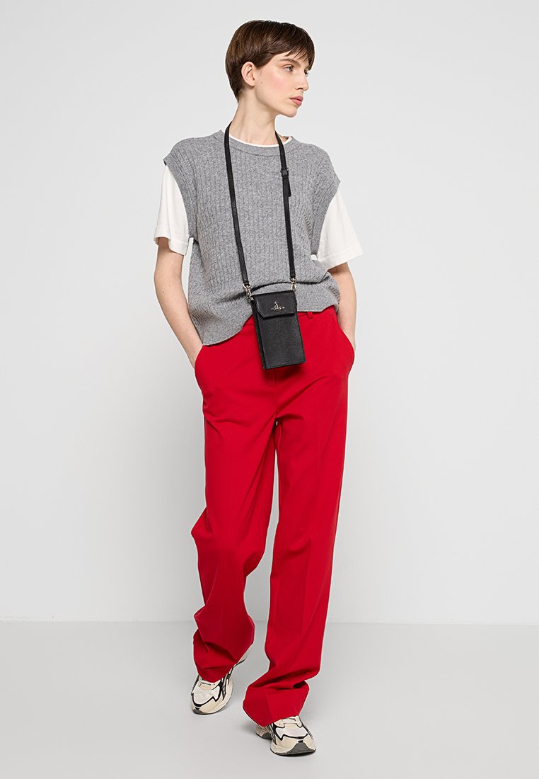 Persona che indossa un gilet di maglia grigio sopra una camicia bianca a maniche corte, pantaloni rossi a gamba larga, sneakers beige e una pochette nera a tracolla per il telefono, in piedi con le mani in tasca.