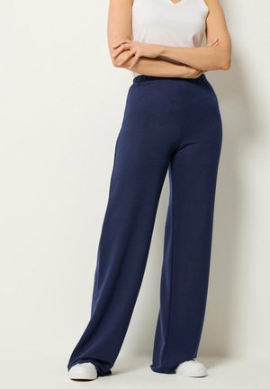 Personne portant un pantalon large bleu marine, des baskets blanches et un haut blanc sans manches, debout les bras croisés devant un fond neutre.