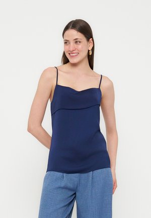 Kvinde smilende iført en navy blå ærmeløs top med tynde stropper og lyseblå bukser med vide ben, stående mod en hvid baggrund.