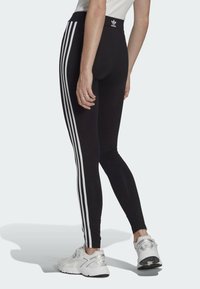 Black athletic leggings with white three-stripe design along the sides. Smooth texture and high waistband. Worn with white sneakers.
