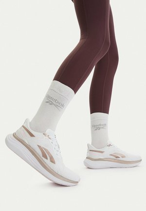 Persona con zapatillas Reebok blancas y beige, calcetines blancos de Reebok y leggings marrones, levantando un pie ligeramente del suelo.