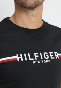 Muž oblečený v černém tričku s kulatým výstřihem s textem "HILFIGER NEW YORK" a červenými a bílými horizontálními pruhy přes hruď.