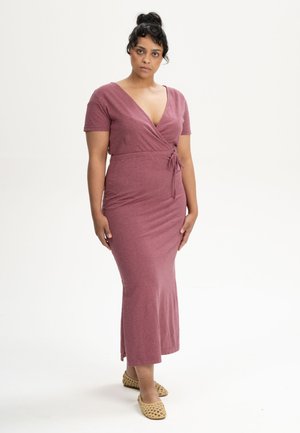 Vrouw met donker haar in een knot, gekleed in een mauve overslagjurk en beige geweven platte schoenen, staand tegen een eenvoudige lichte achtergrond.