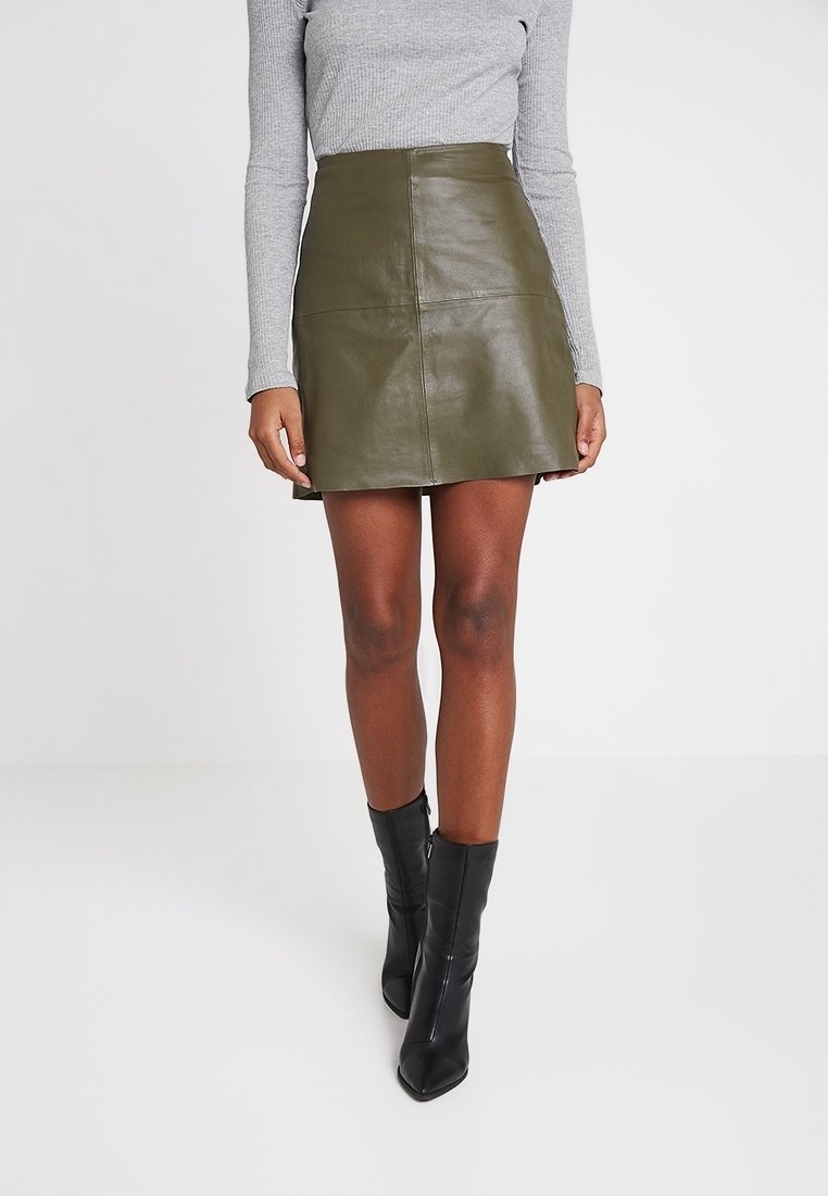 Frau trägt einen olivgrünen Minirock aus Leder, ein graues Langarmshirt und schwarze Stiefeletten, steht vor einem schlichten weißen Hintergrund.