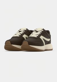 Καφέ αθλητικό παπούτσι με κρεμ κορδόνια και πλαϊνές λεπτομέρειες. Διαθέτει υφασμένο επάνω μέρος, επενδεδυμένη σόλα και χοντρή, ραβδωτή εξωτερική σόλα για πρόσφυση.