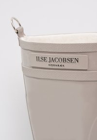 Botte en caoutchouc beige avec une doublure douce, présentant une bande texturée et une plaque logo métallique portant la mention "ILSE JACOBSEN HORNBAEK."