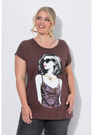 Glimlachende vrouw met blond haar die een bruin T-shirt draagt met een afbeelding van een vrouw met zonnebril en ketting, gecombineerd met een donkere broek.