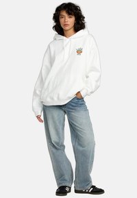 Witte hoodie met een kangoeroezak, voorzien van een kleine groene plantafbeelding en logo op de borst. Gecombineerd met baggy blauwe jeans en zwarte sneakers.