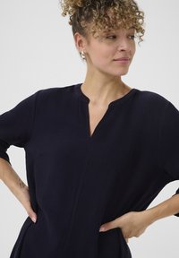 Námořnická modrá tunika s výstřihem do V, ¾ rukávy a dvě boční kapsy. Texturovaná tkanina s uvolněným střihem a jemným prošíváním.