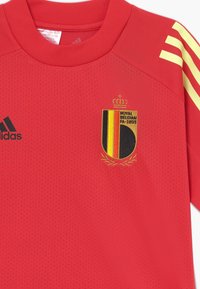 Červený atletický dres vyrobený z prodyšného materiálu, s žlutými akcenty, perforovanou strukturou a vyšitým národním emblémem Belgie.