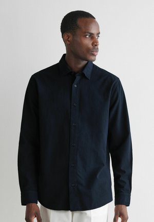 Mann trägt ein schwarzes, langärmliges Hemd mit Knöpfen und weiße Hosen, sieht nach rechts vor einem schlichten hellen Hintergrund.