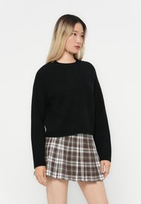 Maglione nero accorciato con maniche lunghe e scollatura rotonda abbinato a una gonna a quadrati marrone e bianca con pieghe e forma svasata.