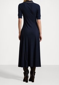 Marinblå klänning i knälängd med korta ärmar, åtsittande liv, utställd kjol och en krage med kontrasterande kant. Slät textur.