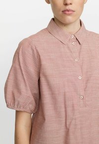 Kurzärmliges, kragenähnliches Hemd in zartem Rosa mit dünnen weißen Streifen. Es hat eine durchgehende Knopfleiste und geraffte Puffärmel für Struktur.
