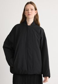 Zwarte pufferjack met een hoge kraag, oversized pasvorm, elastische zoom en geen zichtbare sluitingen. Lichtgewicht stof met een gladde textuur.