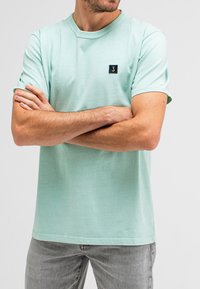 Mint green short-sleeve t-shirt made of soft fabric, featuring a square black logo patch on the upper left chest.