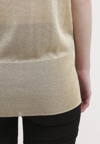 Ljus guldfärgad metallisk stickad topp med ribbad nederkant, med en genomskinlig textur och lös passform, kombinerad med figurnära svarta byxor.
