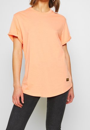 Kvinde iført en løs, kortærmet ferskenfarvet t-shirt og mørkegrå jeans, stående med den ene hånd afslappet ved siden.