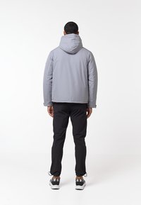 Grå hættejakke med en glat tekstur, parret med sorte bukser. Synlig snøre ved buksebenene, båret med grå sneakers.