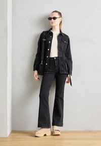 Zwarte denim jacket met zilveren knopen, gecombineerd met hoge zwarte jeans. Model draagt beige platform sandalen en zwarte zonnebril.