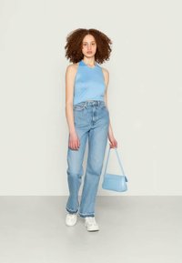 Top sem mangas azul claro, jeans azul desbotado de cintura alta, ténis brancos e uma pequena mala azul com textura suave e forma arredondada.