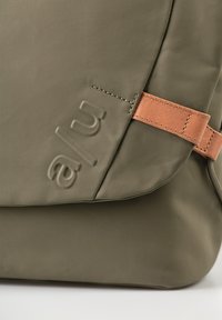 Zaino impermeabile verde oliva con finitura strutturata, dotato di tracolla in pelle color cuoio, logo impresso, forma angolare e dettagli cuciti.