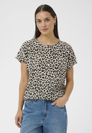 Žena s dlhými tmavými vlasmi, oblečená v krátkom tričku s leopardím vzorom a modrých džínsach, stojaca s jednou rukou v vrecku na jednoliatom pozadí.