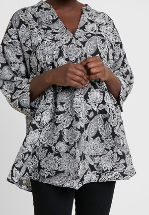 Person trägt eine lockere schwarz-weiß geblümte Paisley-Bluse mit langen Ärmeln und schwarze Hose, Hände vor der Taille verschränkt.