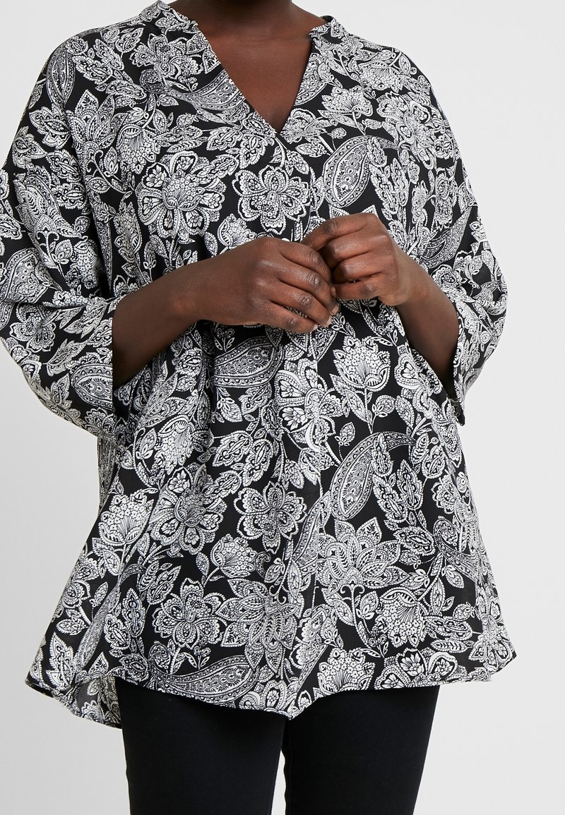 Personne portant un chemisier ample noir et blanc à motif floral et paisley, manches longues, et un pantalon noir, mains jointes à la taille.