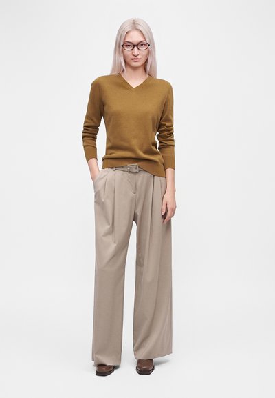 Femme aux longs cheveux blonds et portant des lunettes, vêtue d'un pull marron à col en V, d'un pantalon beige large et de chaussures marron, debout avec une main dans la poche.