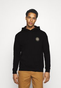 Black hoodie made of soft fabric, featuring a front pocket, drawstring hood, and gold emblem design on the chest.