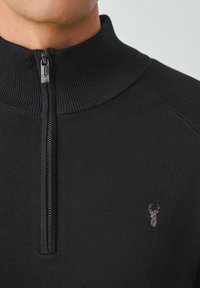 Schwarzer Pullover mit Reißverschluss und geripptem Kragen. Verfügt über ein kleines gesticktes Reh-Logo auf der Brust und ein strukturiertes Strickmuster.