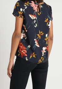 Blusa floral de tela azul marino con mangas cortas, que presenta un detalle de abertura en la espalda y un patrón variado de flores rojas, rosas y amarillas.